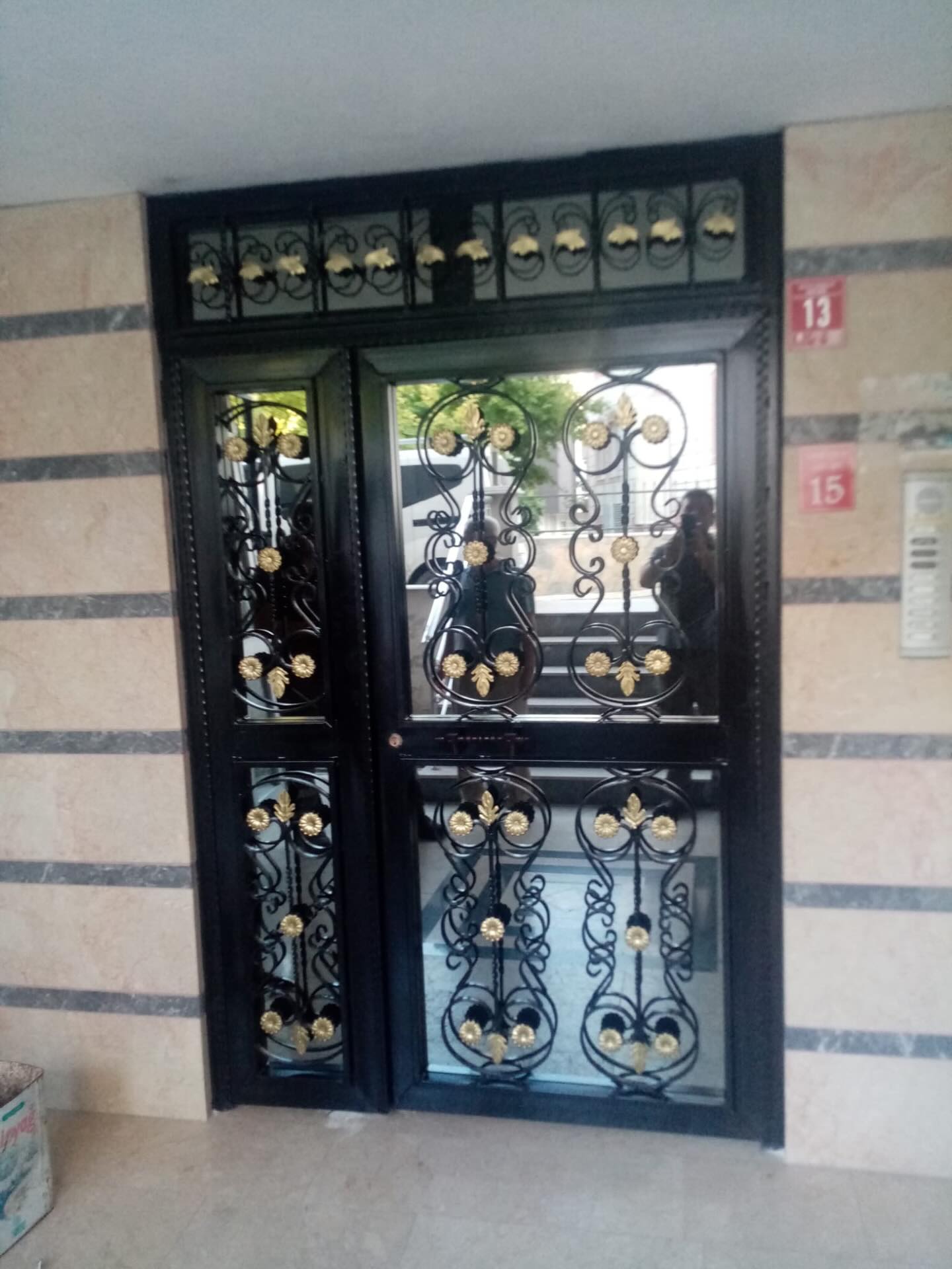 Apartman girişinde siyah ferforje çerçeveli, camlı ve altın çiçek motifli çift kanatlı kapı