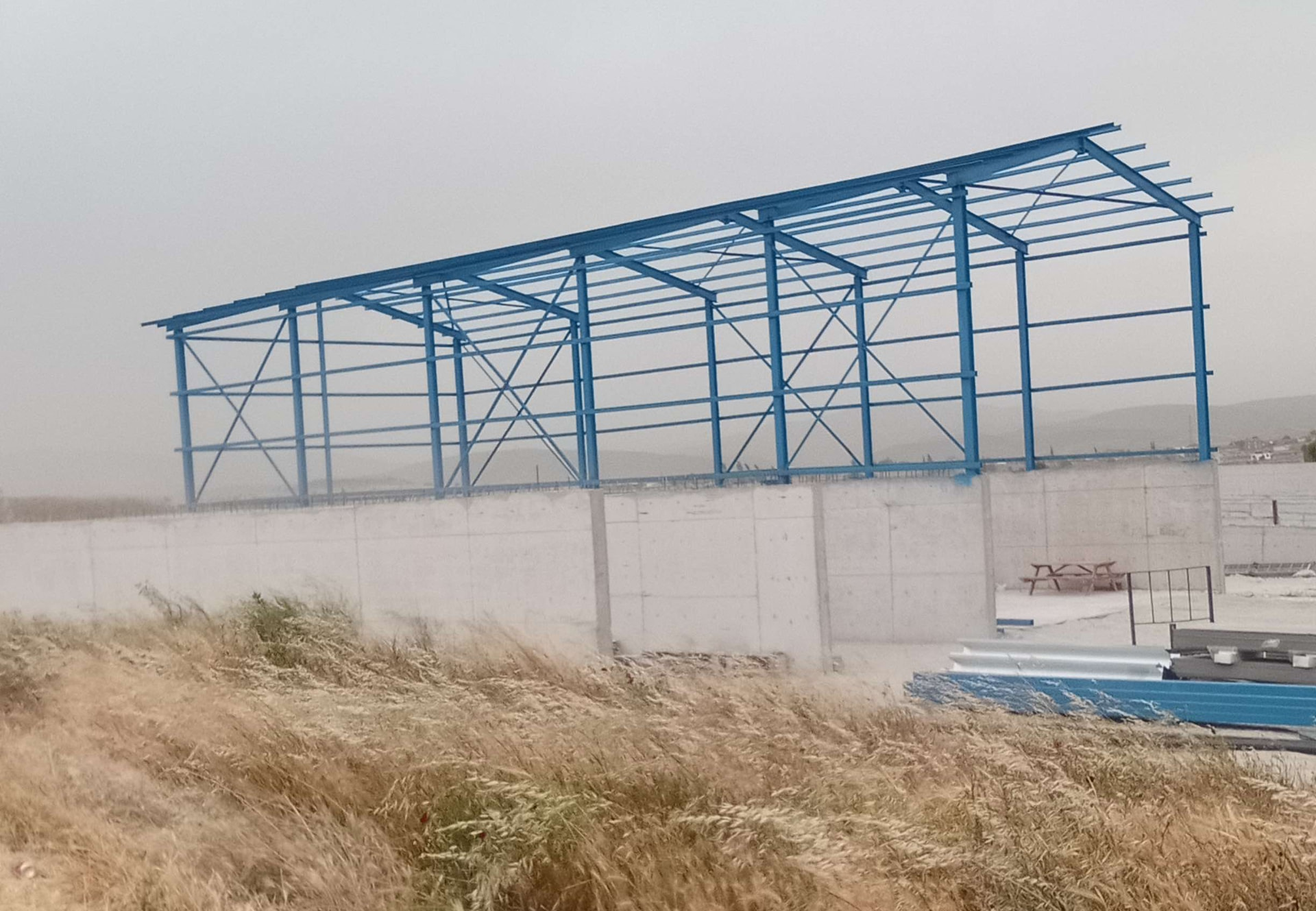 Yüksek beton duvar üzerinde mavi kolonlar, kirişler ve çaprazlardan oluşan çelik depo iskeleti