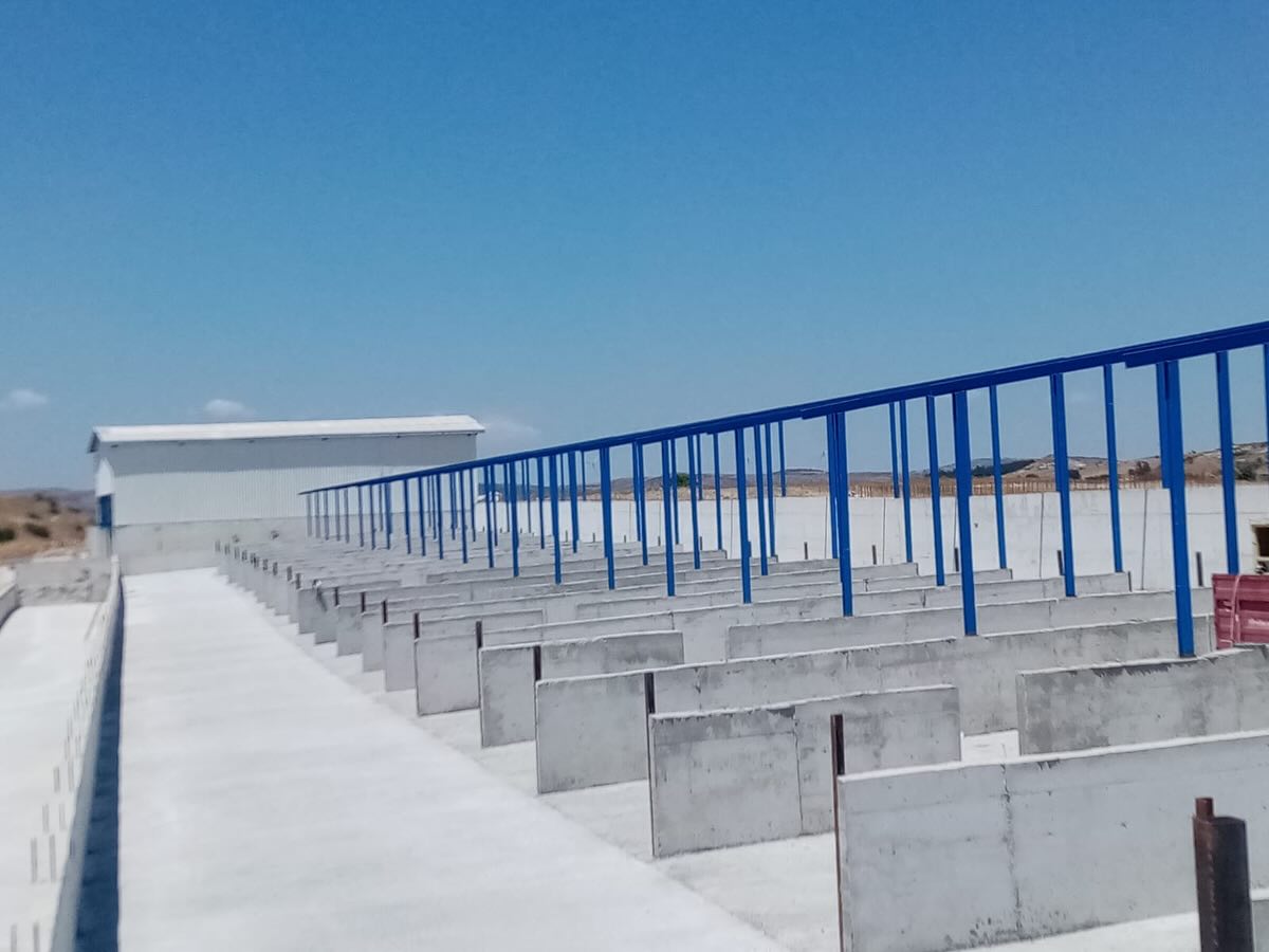 Beton bölmeli ağıl üzerinde sıralanmış mavi çelik korkuluklar ve uzaktaki depo yapısı