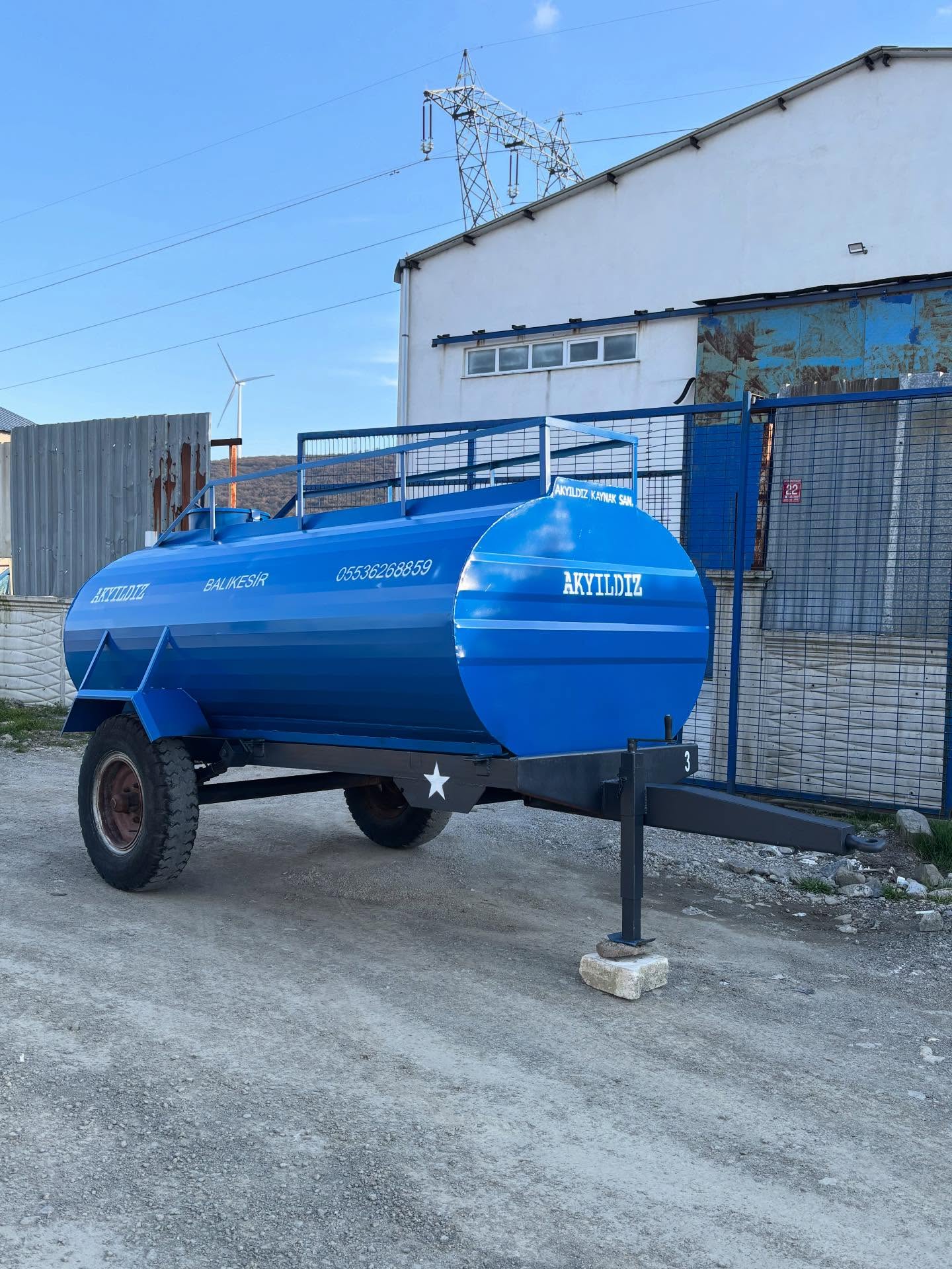Mavi boyalı, üstünde tel korkuluk bulunan tek dingilli çekilir su tankerinin yandan görünümü